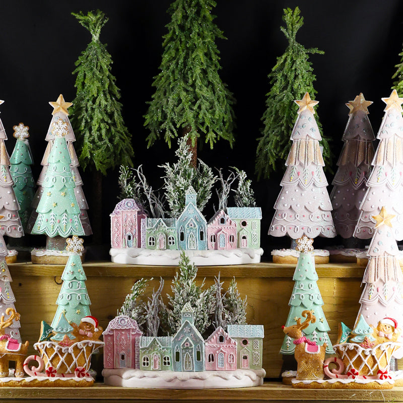 A collection of hand-painted Christmas village figurines and miniature trees displayed on wooden shelves against a black background. The set includes pastel-colored houses in pink, blue, and green, decorated Christmas trees, and gingerbread-themed sleighs with cookies.