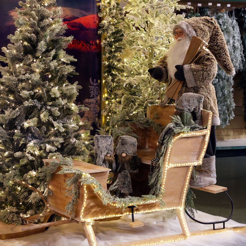 A festive Christmas display featuring a Santa Claus figure in a fur-trimmed suit standing beside a wooden sleigh decorated with warm white string lights, evergreen garlands, and faux snow, with illuminated Christmas trees in the background.