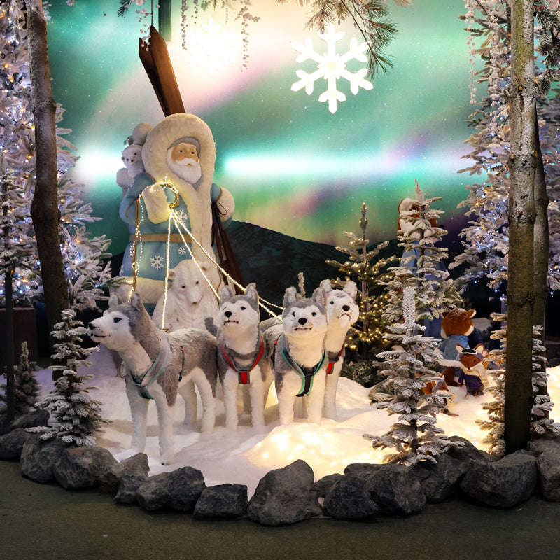 A festive Santa Claus and dog-sled display in a snowy forest setting. Santa Claus stands on a sleigh pulled by four husky dogs, with additional snow-covered trees, rocks, and a glowing aurora backdrop.