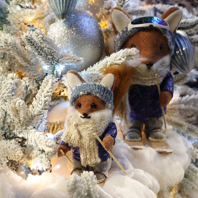 Two plush toy foxes dressed in winter ski outfits, posed among flocked pine branches and silver ornaments on a snowy white surface with warm string lights.
