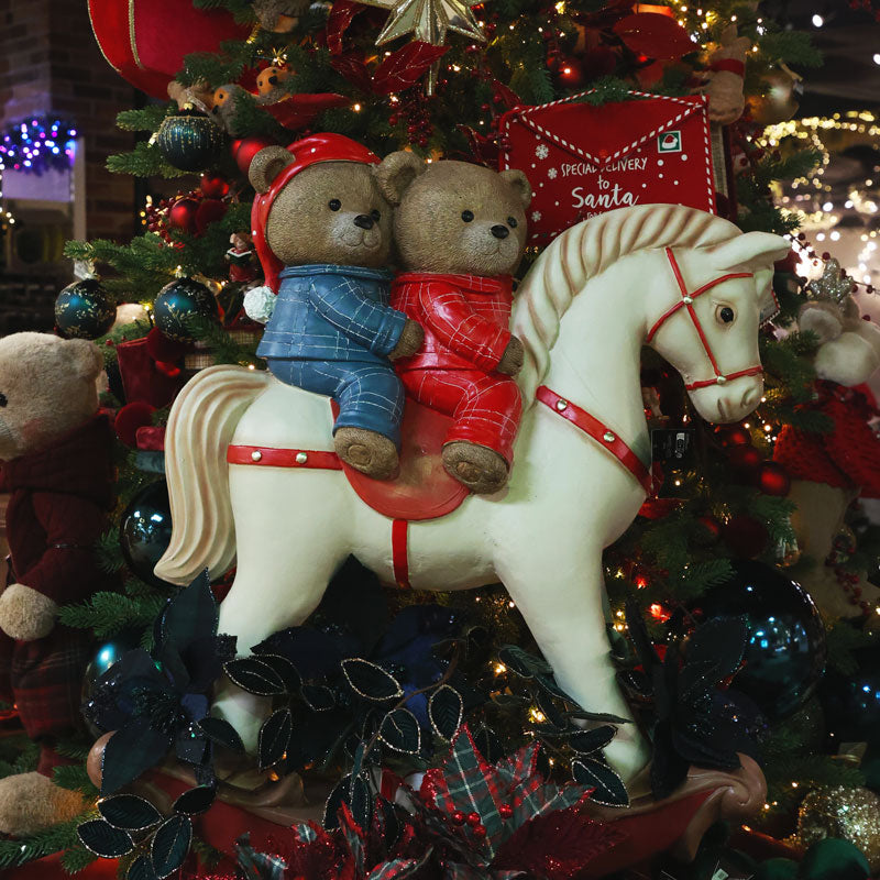 A white rocking horse Christmas ornament decorated with red harness details, carrying two teddy bears in blue and red pajamas. The ornament is displayed on a decorated Christmas tree with lights, ornaments, and other holiday decorations.