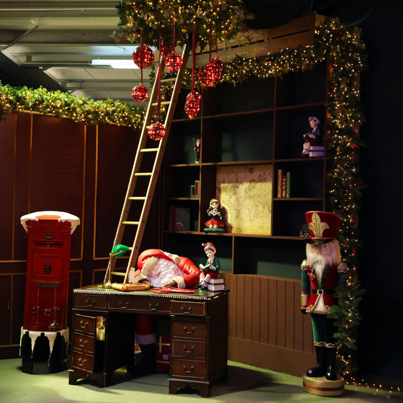 A festive holiday-themed room scene with a wooden desk, ladder, red hanging ornaments, Christmas garland, bookshelves, a red post box, and a nutcracker figure. A person in a Santa outfit is lying on the desk, creating a surreal, staged scene.
