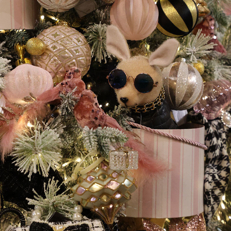 A plush dog toy wearing round black sunglasses and a black collar with a gold chain, nestled among a decorated Christmas tree with pink ornaments, white and gold baubles, lights, and a striped gift box.