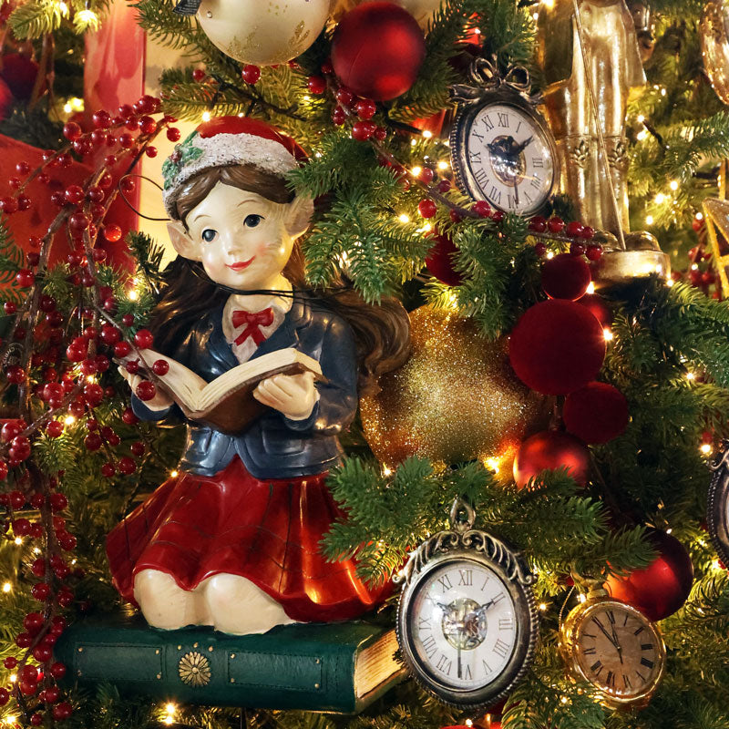 A Christmas-themed decorative figurine of a girl with pointed ears reading a book, posed on a green book among a decorated Christmas tree with red ornaments, gold baubles, and antique-style clocks.