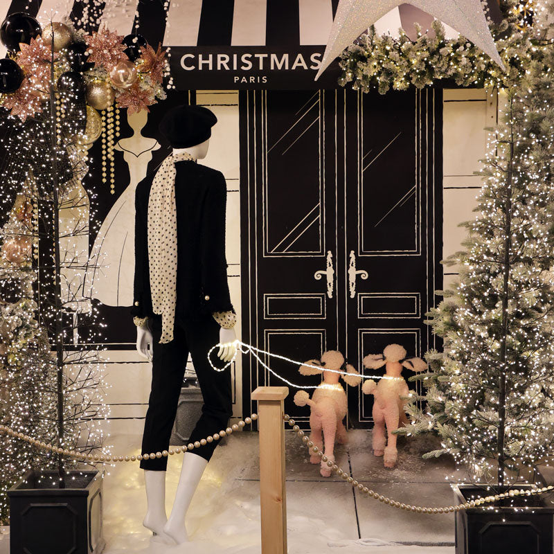 A festive Christmas storefront display with a mannequin dressed in a black outfit and patterned scarf, black double doors, decorated trees with lights, and two plush poodle figures in the foreground.