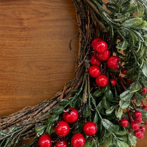 An artificial Christmas wreath made of rustic brown grapevine branches, decorated with glossy green leaves and bright red berries, shown on a warm brown wooden background.
