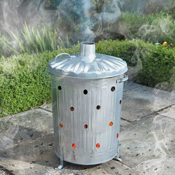 A metal garden bin with a central chimney and air ventilation holes, sat on a garden patio. The bin contains burning waste with smoke coming out of the chimney.