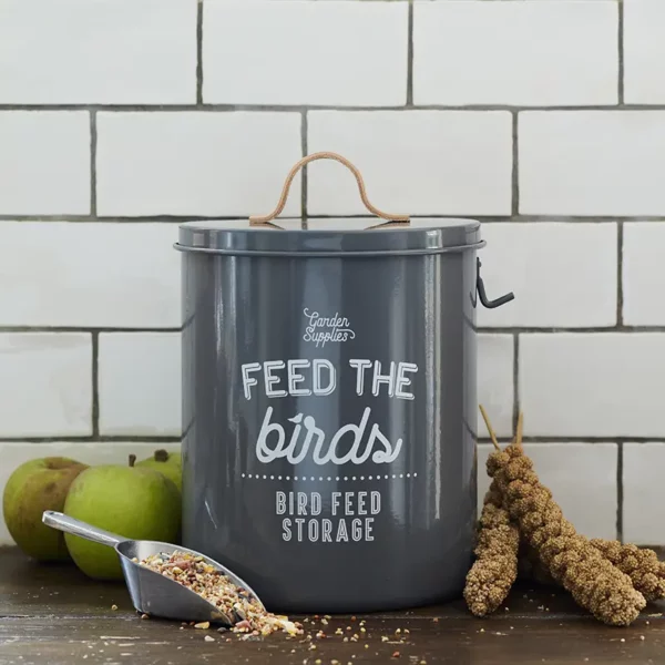 A Burgon & Ball 'Feed the Birds' Bird Food Tin sat on a kitchen counter with a scoop of feed. The tin is charcoal grey coloured with white typography.