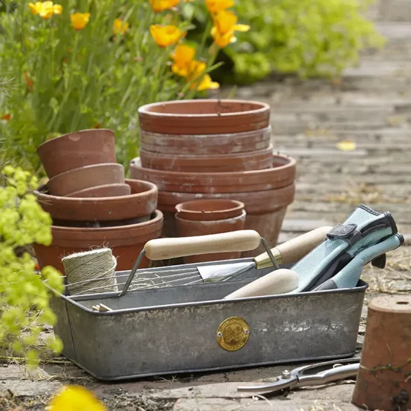A metal Burgon & Ball Sophie Conran Galvanized Trug placed on a path holding a range of gardening tools and gloves.