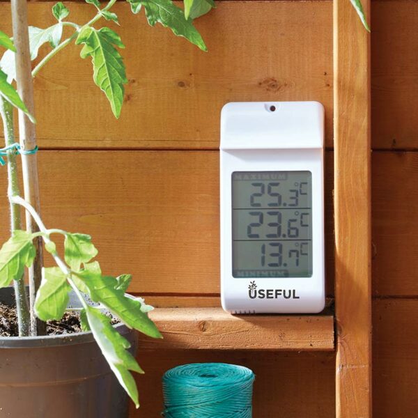 A white digital thermometer resting on a wooden shelf next to a plant. The thermometer screen has three sections for current, maximum and minimum temperature.