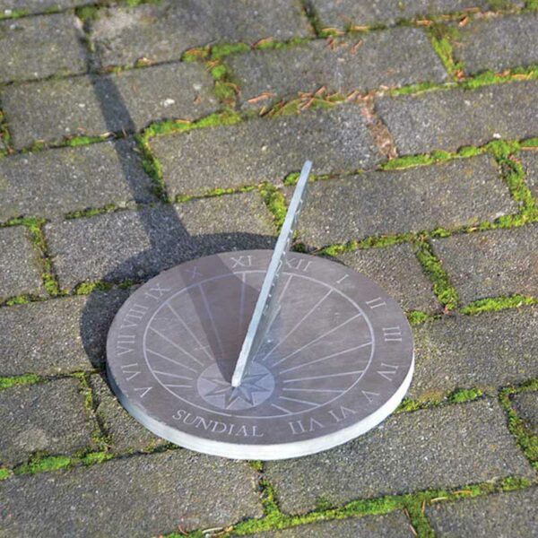 A round slate sundial sat on a mossy patio. The sundial is reading about 08:45 AM.