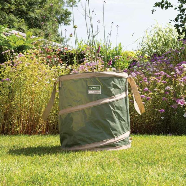 A small green and cream trimmed pop-up gardening tidy bag for collecting garden waste., sat outside on a lawn. It has cream handles and a spring that provides the shape.